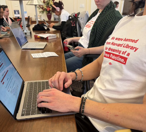 IMG_4462 Student protestors wearing white shirts with red text stating "We were banned from Harvard Library for dreaming of a Free Palestine" working on their laptops.