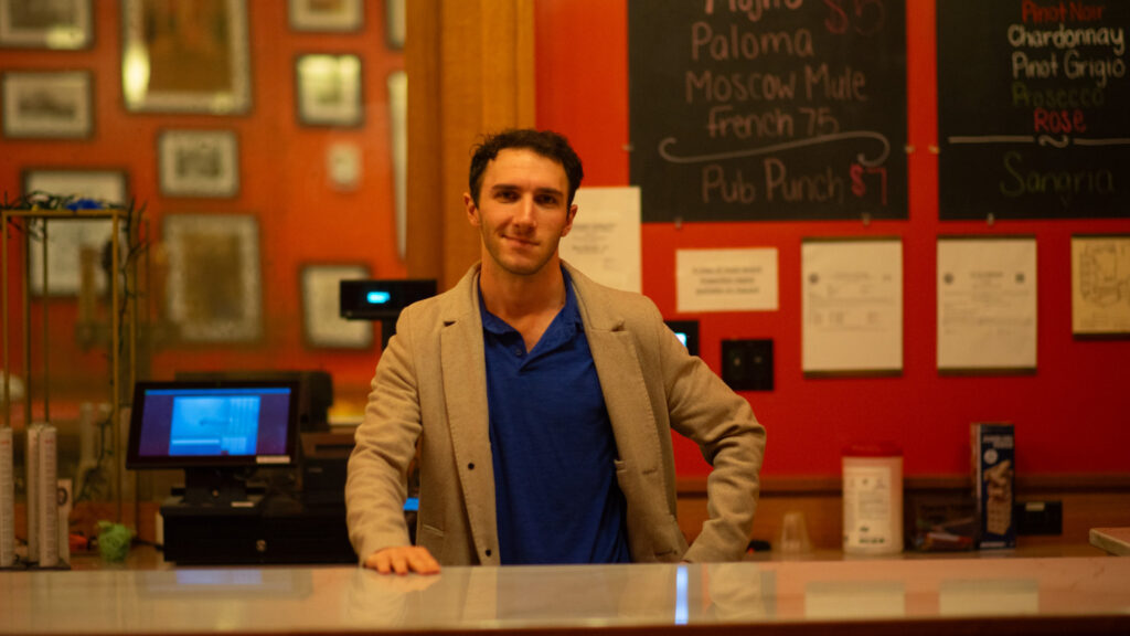 A man (James Atlas, HLS '25) stands behind the table at the HLS Pub.