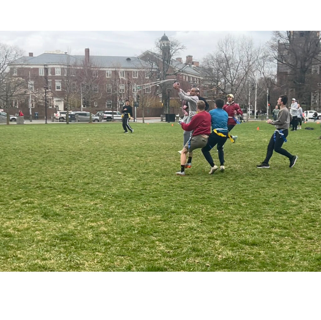 Several flag football players running in Cambridge Common park in the midst of a match. A well-timed throw from Manu Iribane (all black, standing in the background) leads to a contested catch by Salim Sleiman (in the foreground in gray, amidst several other players) , LLM '25, makes a contested catch midfield.