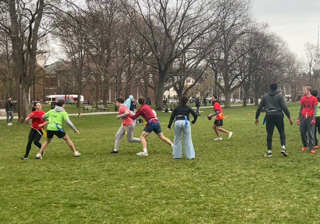 Several flag football players running in Cambridge Common park in the midst of a match. Parth Patel, HLS '27 (orange shirt, white shoes), prepares to throw.