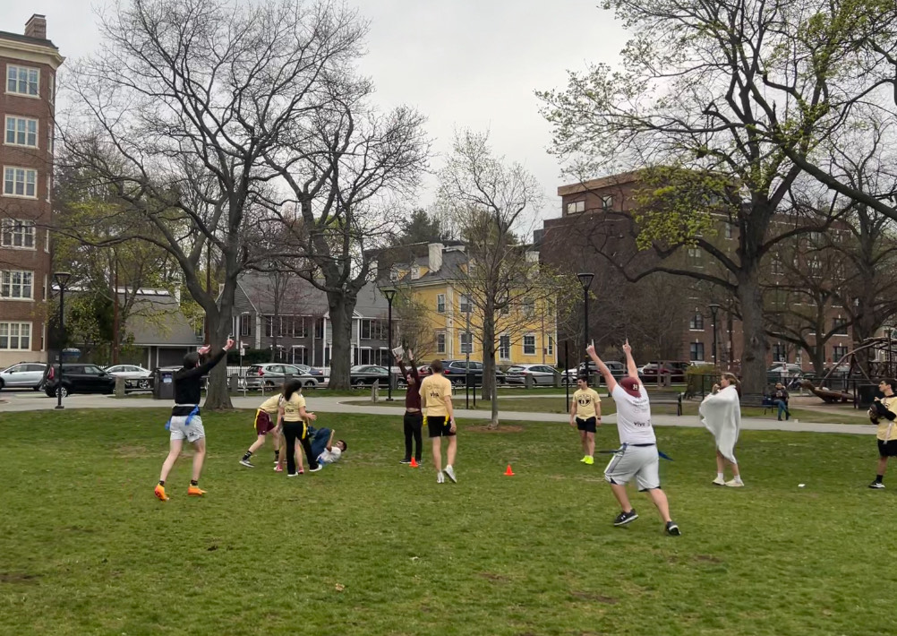Several flag football players running in Cambridge Common park in the midst of a match. A well-placed throw from Brian Broderick(in white shirt and pants,, standing in the foreground) leads to a contested touchdown catch by Aryan Agrawal (on the floor in the background, amidst several other players) , LLM '25.