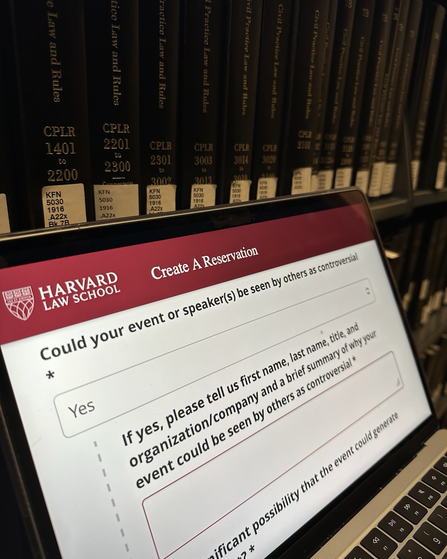 A laptop open to Harvard Law School’s event reservation form, with “Yes” selected for whether the event or speakers could be seen as controversial, in front of shelves of law books.